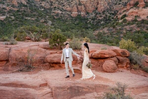 Elopement Wedding A couple walks hand in hand on red rock formations in a desert landscape, surrounded by green shrubs and rocky cliffs. The man wears a light suit, and the woman is in an ivory dress, holding a bouquet. Elopements Inc