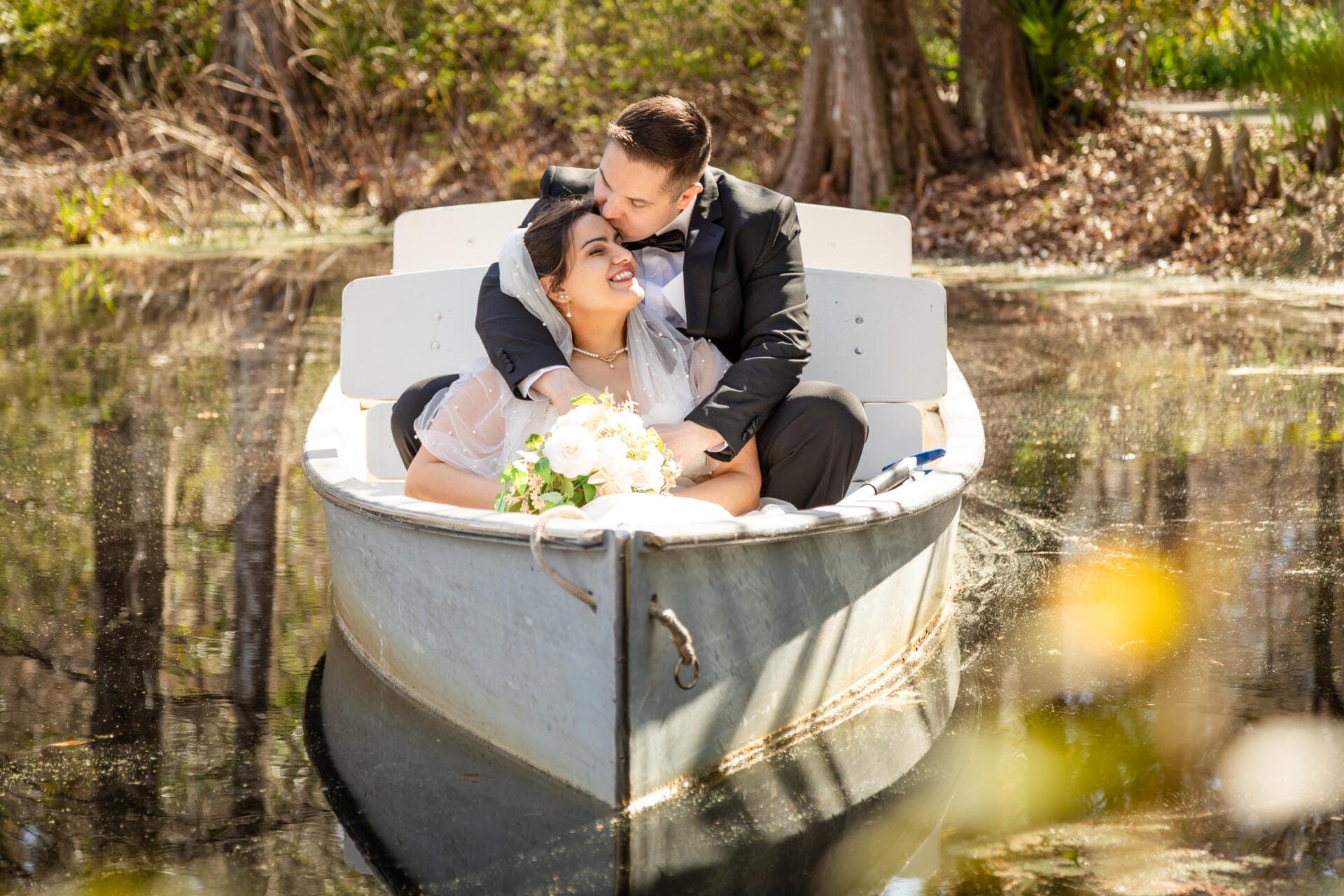 Elopement Packages with A couple celebrating their elopement drifts together in a quaint rowboat across a tranquil pond. The partner in a classic black suit gently kisses the other's forehead; the bride beams, cradling her bouquet while dressed in an elegant white gown and flowing veil. Sunlight dances through the canopy of trees, casting a warm, romantic glow over this intimate wedding moment. by Elopements Inc.